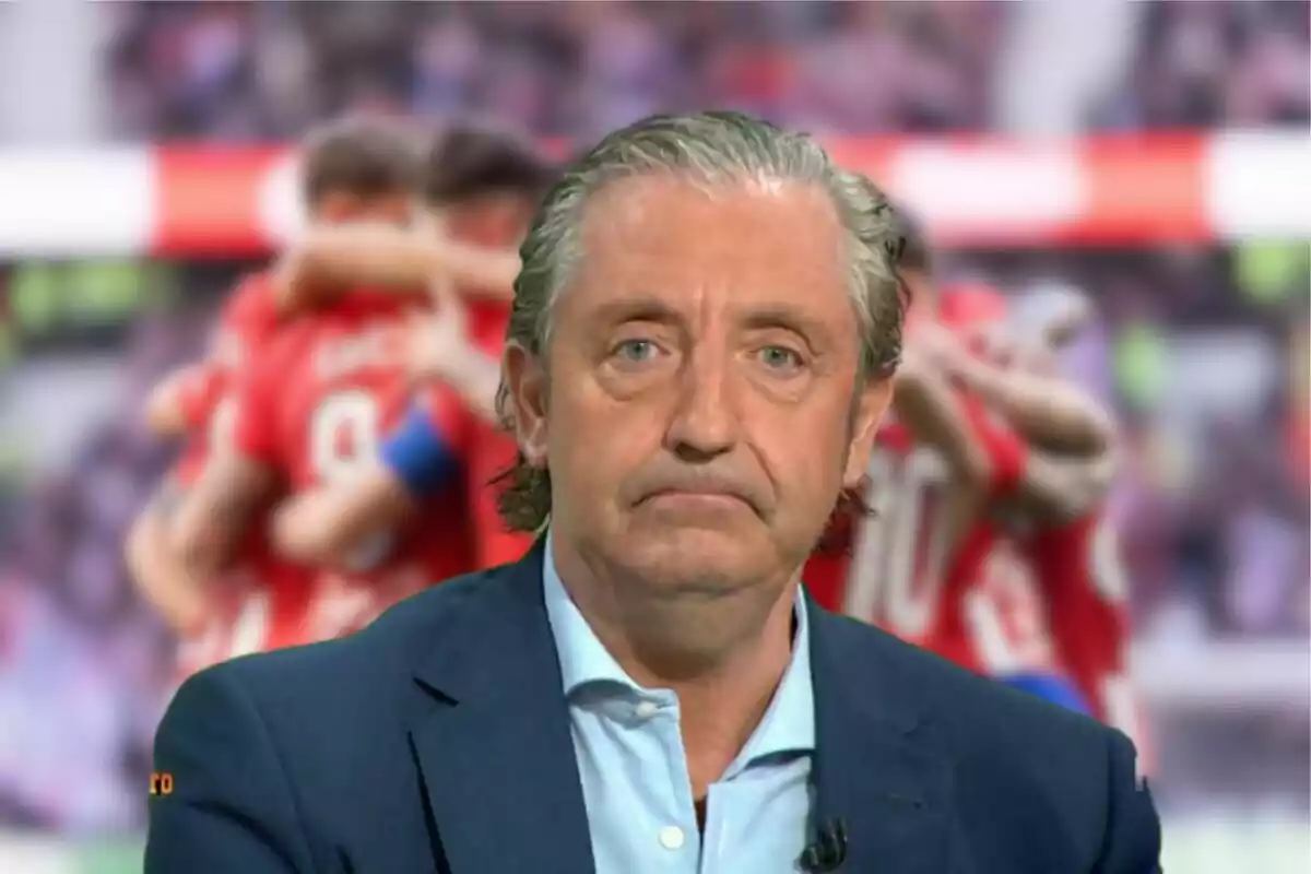 Un hombre de cabello canoso y expresión seria, vestido con un traje azul y camisa clara, aparece en primer plano con un fondo desenfocado de jugadores de fútbol celebrando.