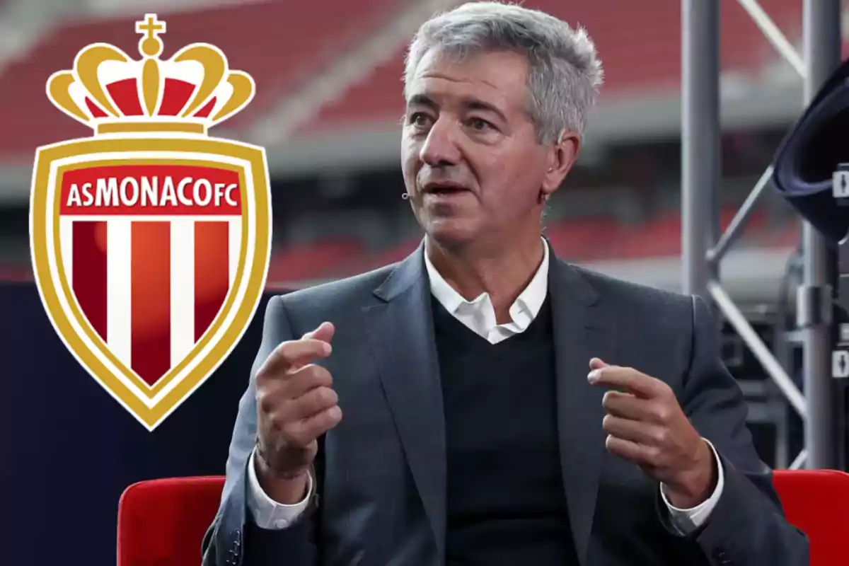 Un hombre de cabello canoso en traje oscuro gesticula mientras habla con el logo del AS Monaco FC al fondo.