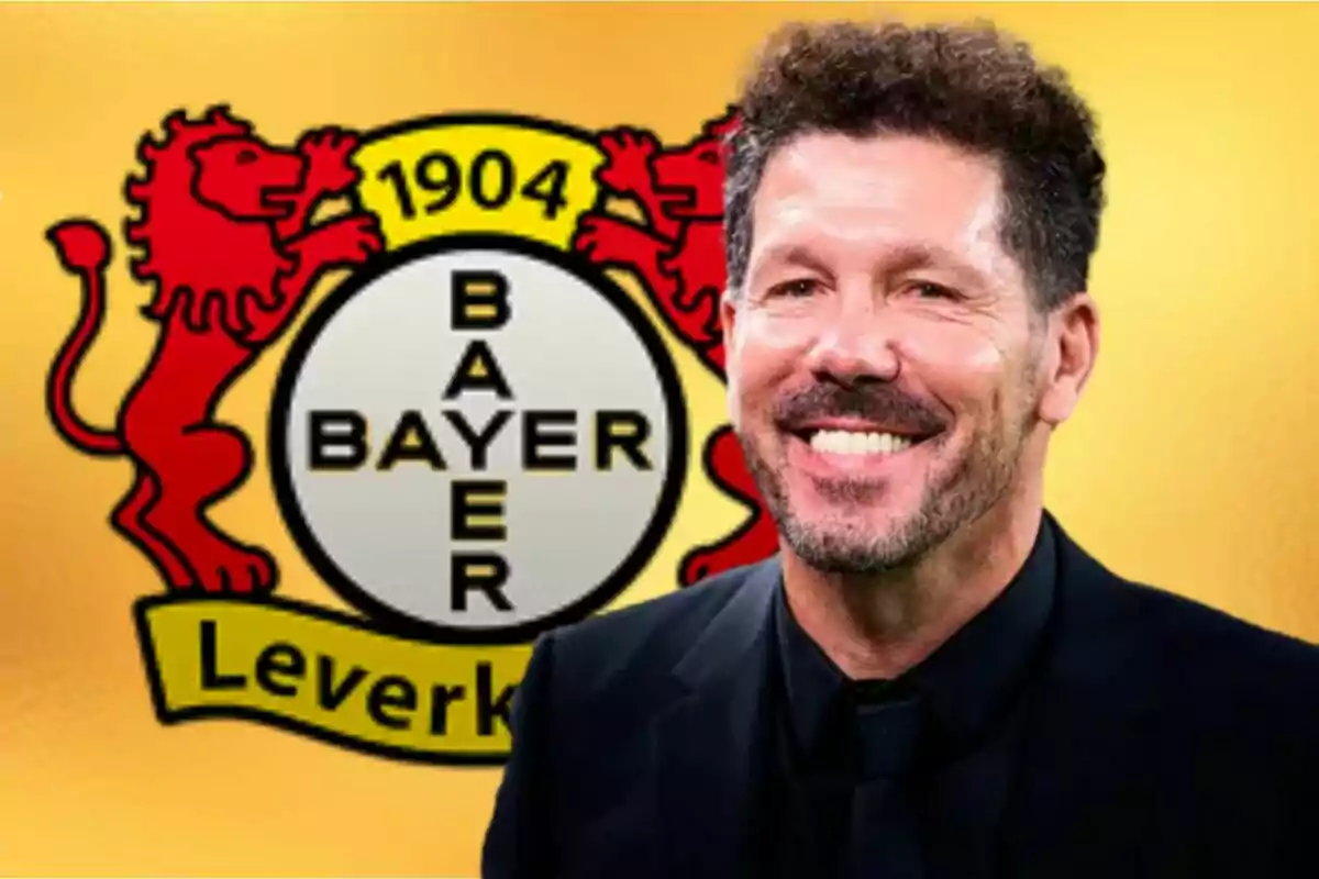 Un hombre sonriente con barba y cabello rizado frente al logo del Bayer Leverkusen.