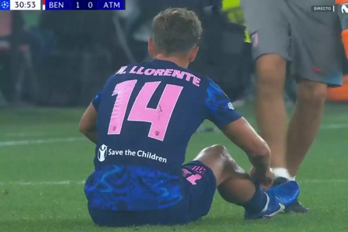 Un jugador de fútbol sentado en el campo durante un partido, con el marcador mostrando BEN 1 - 0 ATM y el dorsal número 14 en su camiseta.