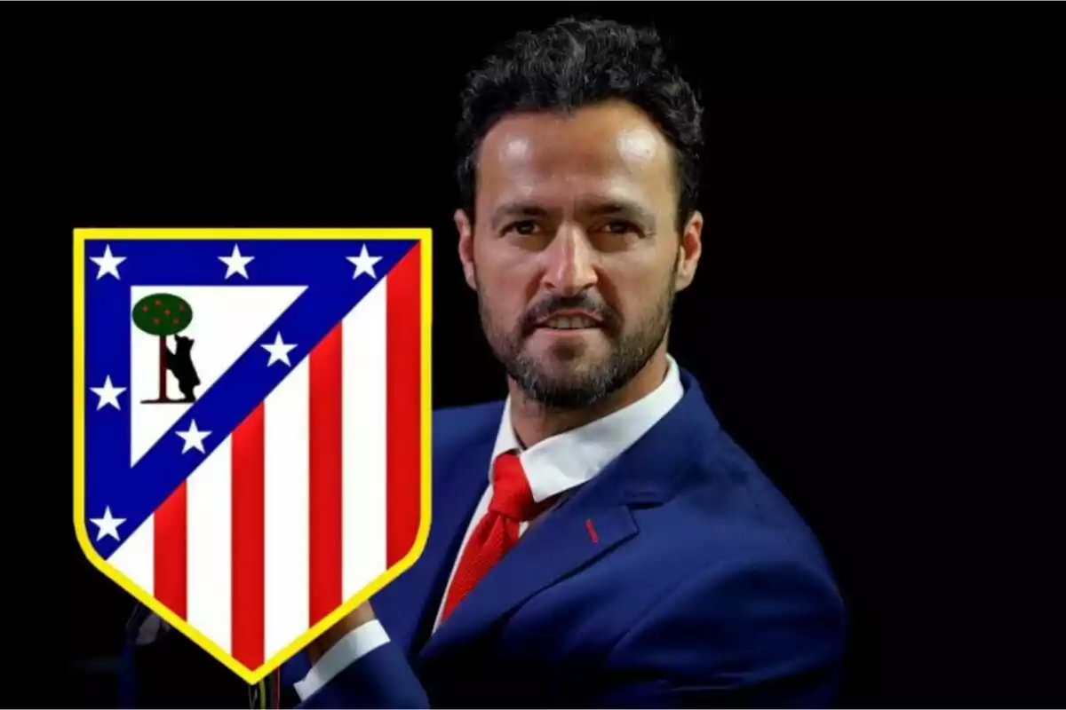 Hombre con traje azul y corbata roja junto al escudo del Atlético de Madrid.