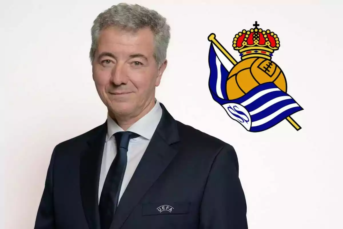 Un hombre con traje y corbata, con el logo de la UEFA en su bolsillo, posando frente a un fondo blanco con el escudo de la Real Sociedad.