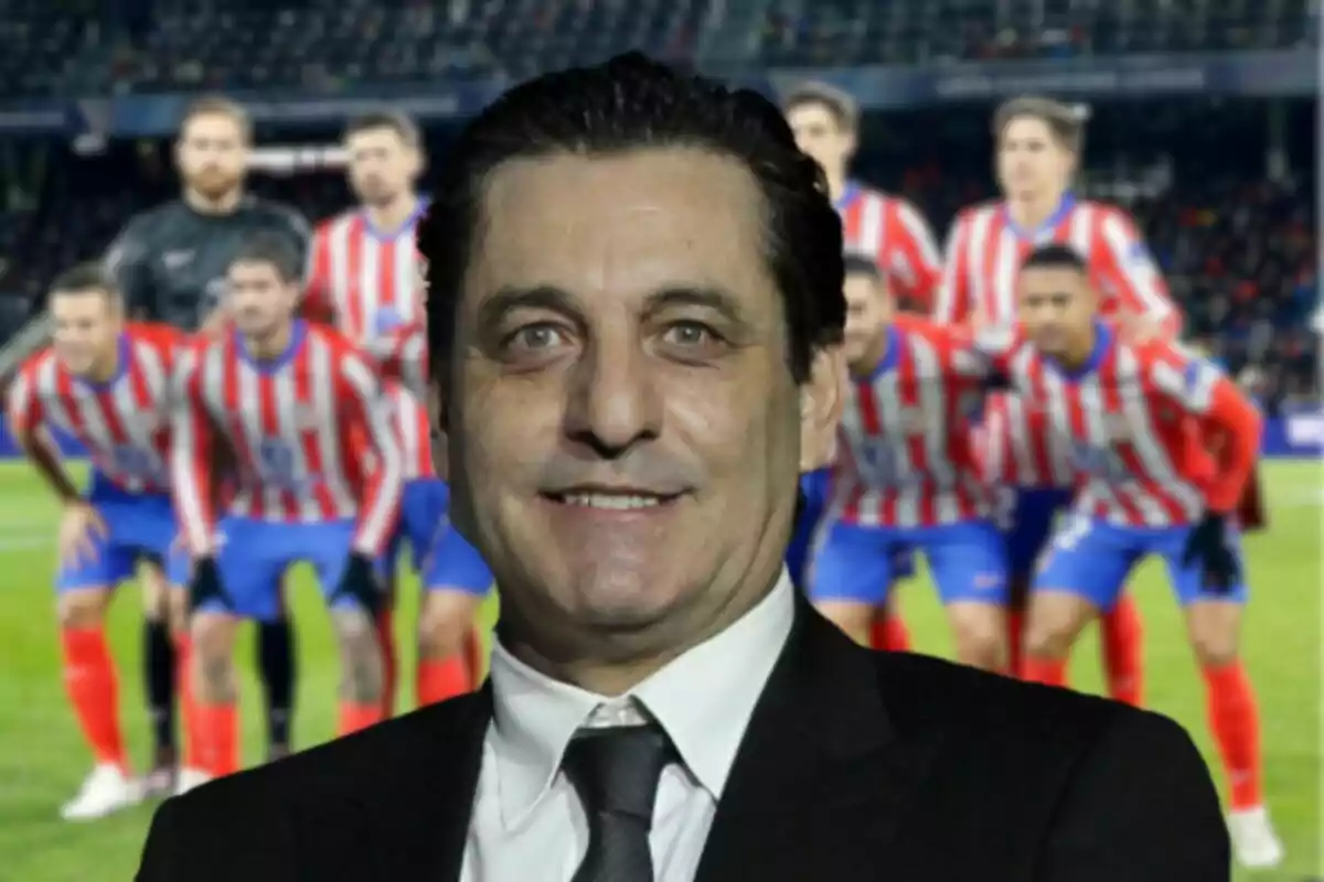 A man in a suit smiles in front of a soccer team in red and white striped uniforms at a stadium.