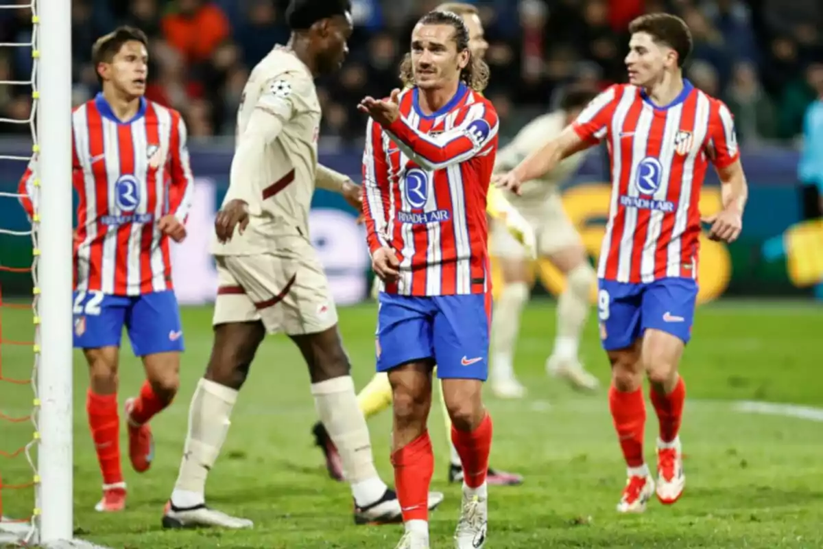 Jugadores de fútbol en el campo durante un partido, uno de ellos con uniforme a rayas rojas y blancas parece estar gesticulando hacia otro jugador con uniforme claro.