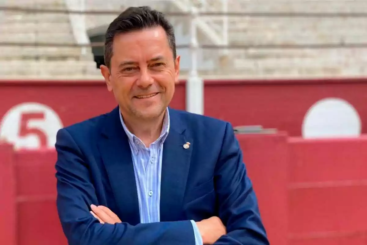 Hombre de traje azul sonriendo con los brazos cruzados frente a una pared roja con el número 5 en el fondo.