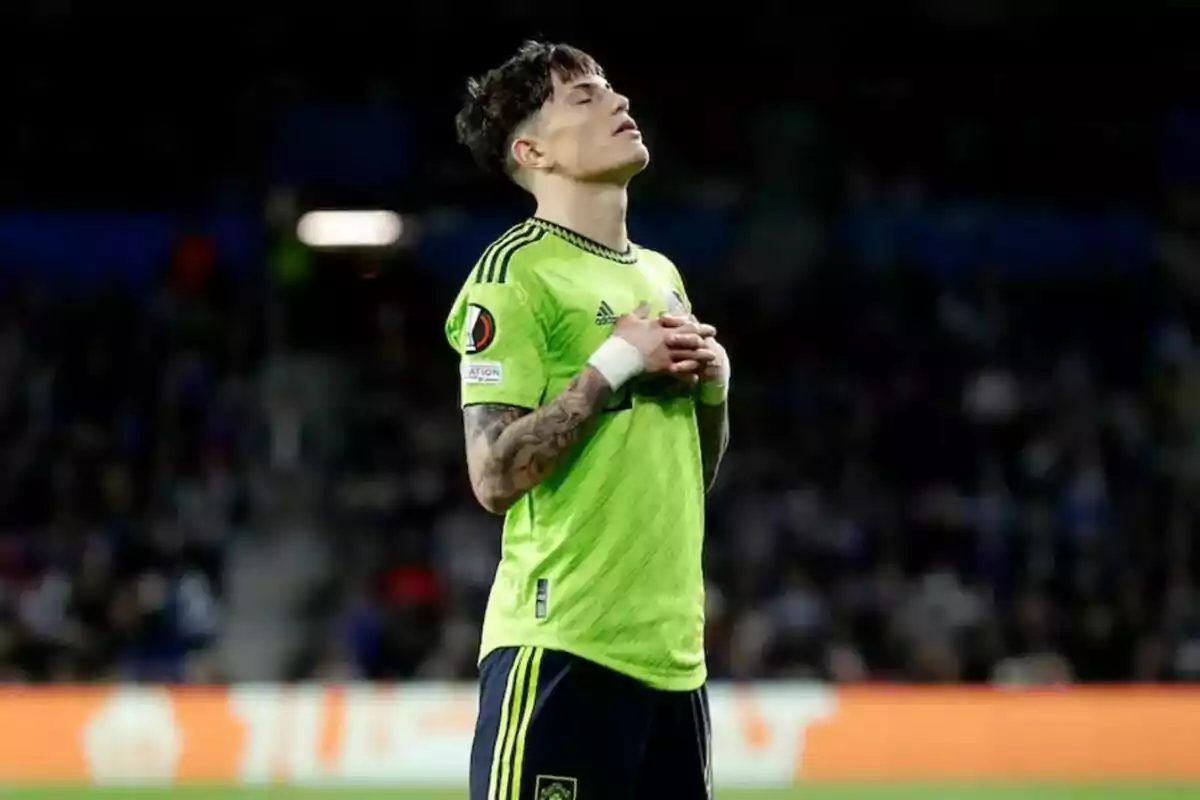 Jugador de fútbol con uniforme verde en el campo, con las manos sobre el pecho y los ojos cerrados.