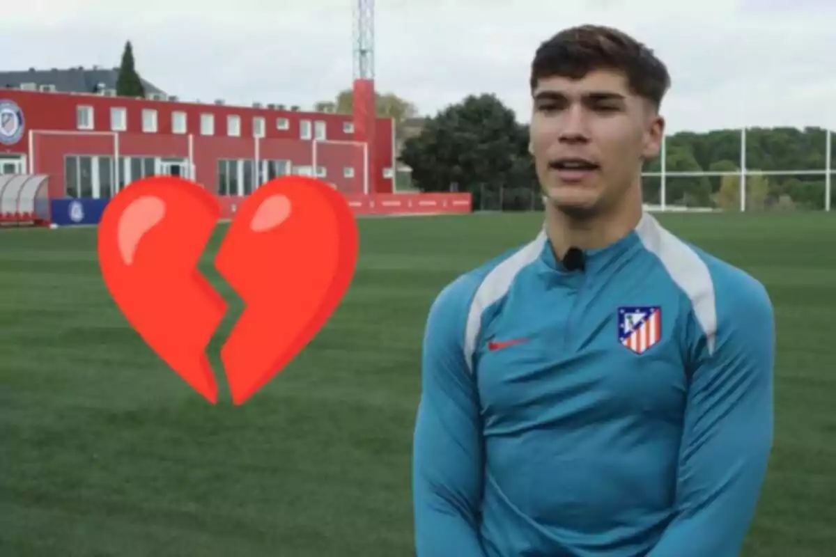 Un jugador de fútbol con uniforme del Atlético de Madrid está de pie en un campo de entrenamiento con un emoji de corazón roto superpuesto.