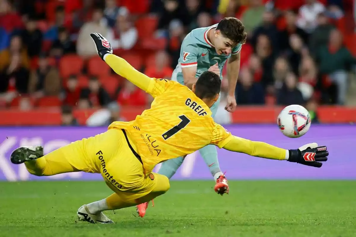 A soccer player attempts to head the ball as the goalkeeper dives to stop it in a stadium full of spectators.