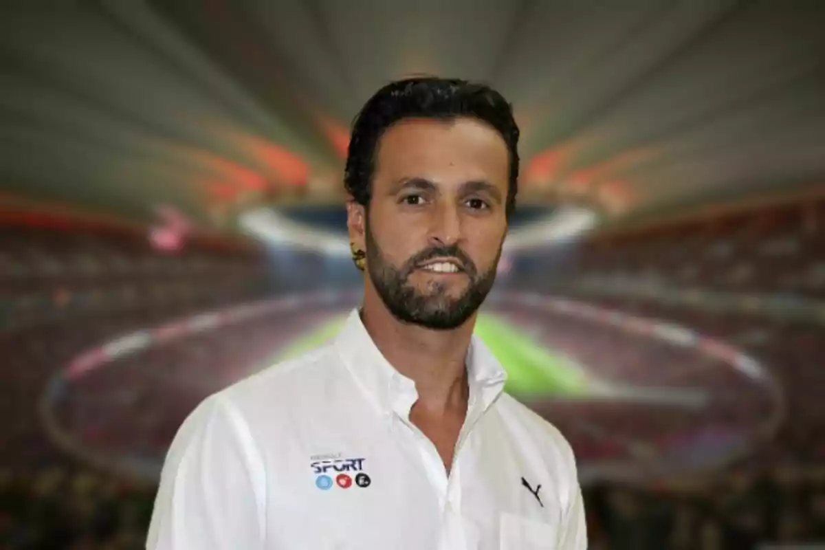 Hombre con barba y camisa blanca posando frente a un estadio desenfocado.