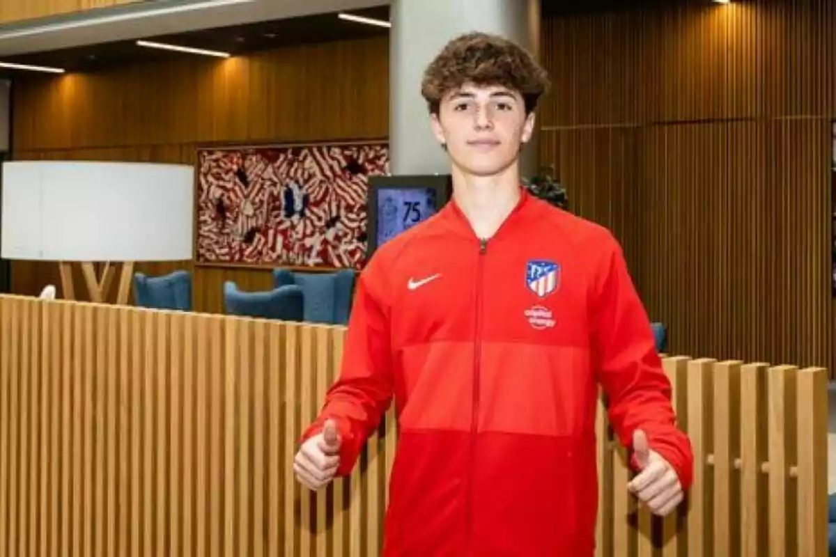 Young man in red Atletico Madrid jacket posing with thumbs up in modern interior with wooden walls and big lamp.