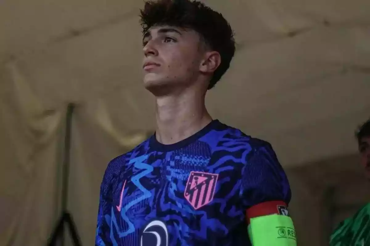 A young footballer in a blue uniform with pink details of Atlético de Madrid, looking up with a serious expression.