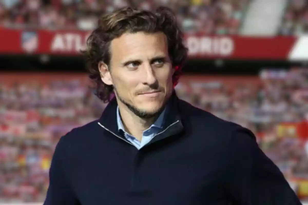 Un hombre con cabello rizado y barba ligera, vestido con un suéter oscuro, está de pie en un estadio de fútbol con un cartel de fondo que dice "ATLETI MADRID".