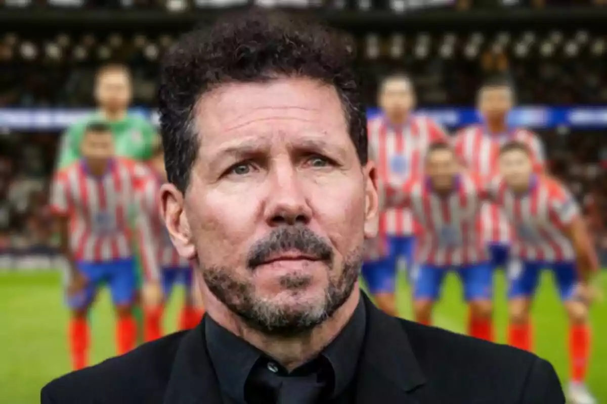 Un hombre con barba y cabello rizado en primer plano con un equipo de fútbol desenfocado al fondo en un estadio.