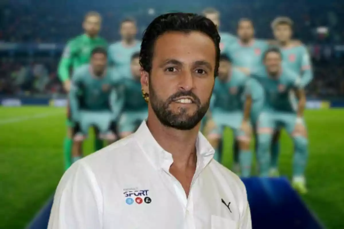 Un hombre con barba y camisa blanca está de pie frente a un grupo de jugadores de fútbol desenfocados en un estadio.