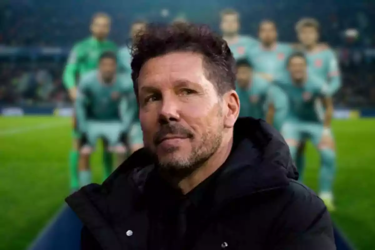 A bearded man with dark hair stands in the foreground, with a group of people out of focus in the background on a soccer field.