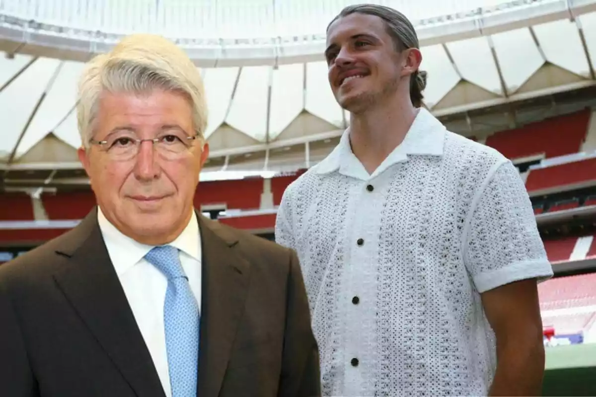 Dos hombres posan en un estadio, uno de ellos lleva traje y corbata, mientras que el otro viste una camisa blanca de punto.