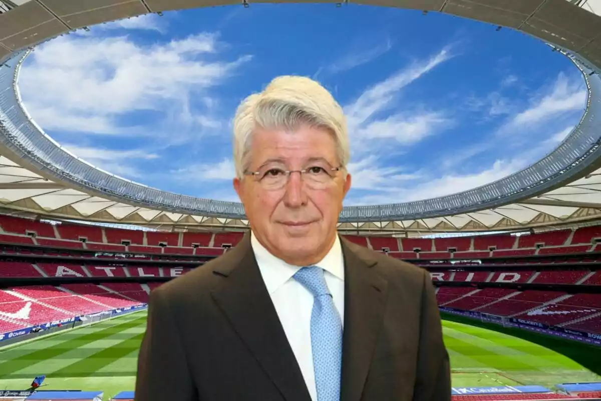 Un hombre de cabello canoso y gafas, vestido con traje y corbata, posando en un estadio de fútbol con gradas rojas y un cielo azul de fondo.