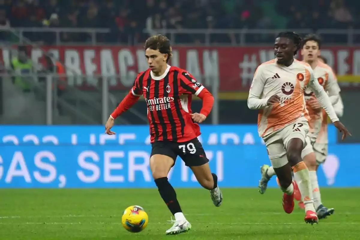 Un jugador de fútbol con uniforme rojo y negro corre con el balón mientras es perseguido por un jugador con uniforme blanco y naranja en un estadio.