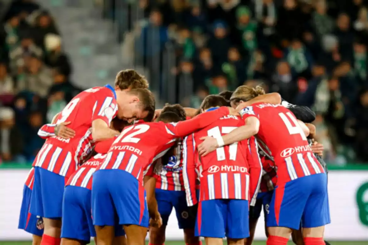 Jugadores de fútbol con uniformes rojos y azules se abrazan en círculo en el campo.