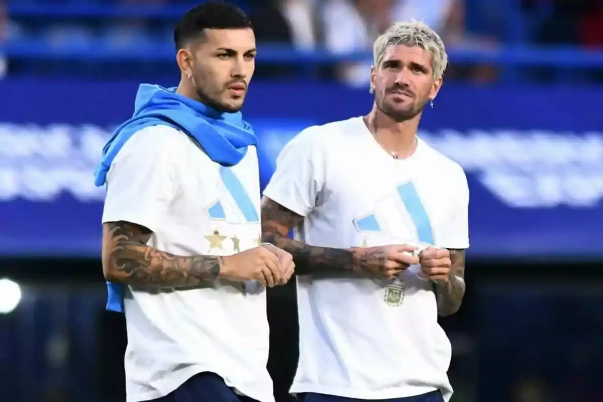 Dos hombres con camisetas blancas y detalles celestes están de pie en un entorno deportivo, uno lleva una sudadera azul sobre los hombros y ambos tienen tatuajes visibles en los brazos.