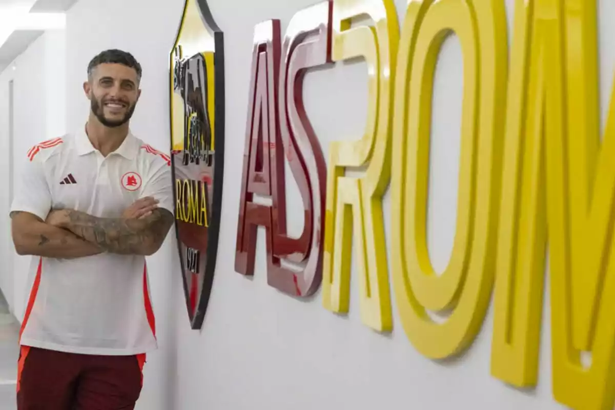 Un hombre con una camiseta blanca y roja está de pie junto a un letrero grande que dice "AS ROMA" en un pasillo iluminado.