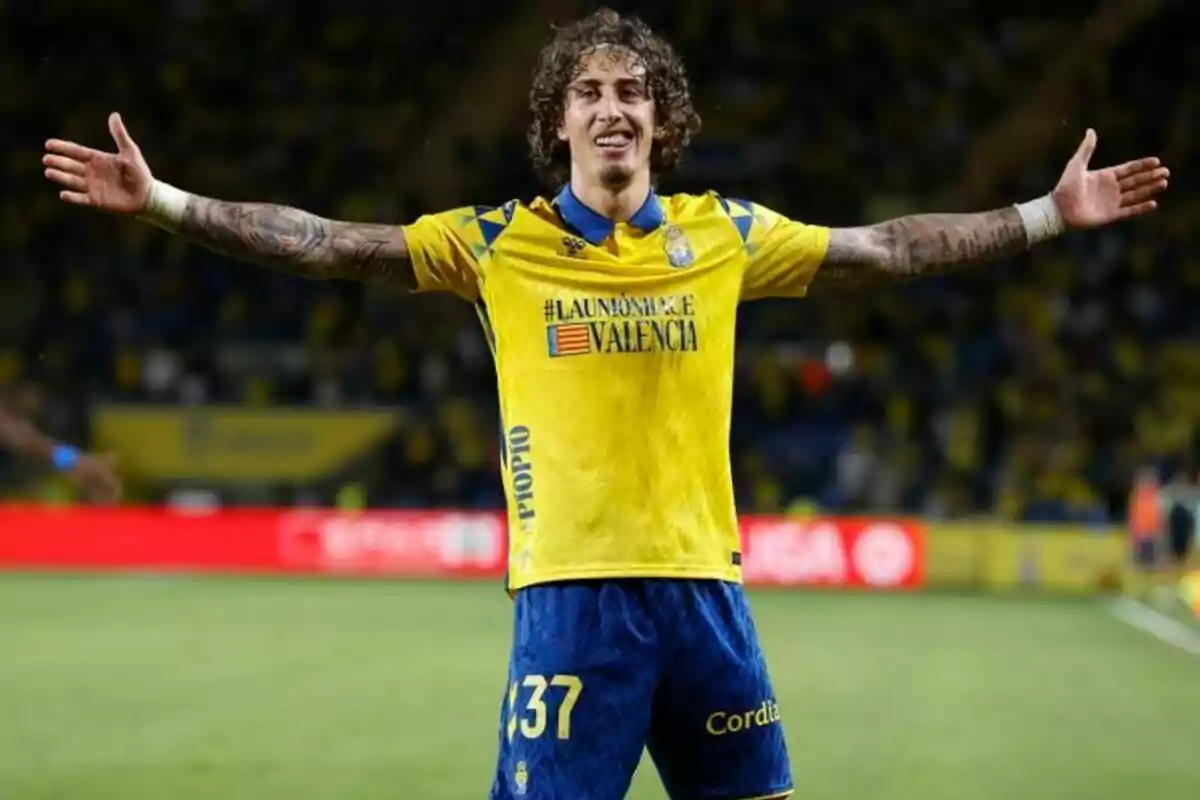 Un jugador de fútbol con uniforme amarillo y azul celebra con los brazos abiertos en un estadio.