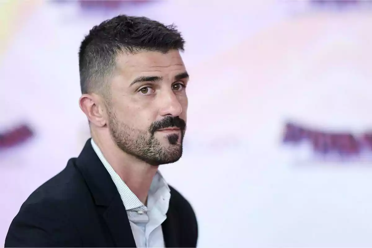 Un hombre con barba y cabello corto, vistiendo un traje oscuro y camisa clara, mira hacia la cámara con una expresión seria, sobre un fondo desenfocado.