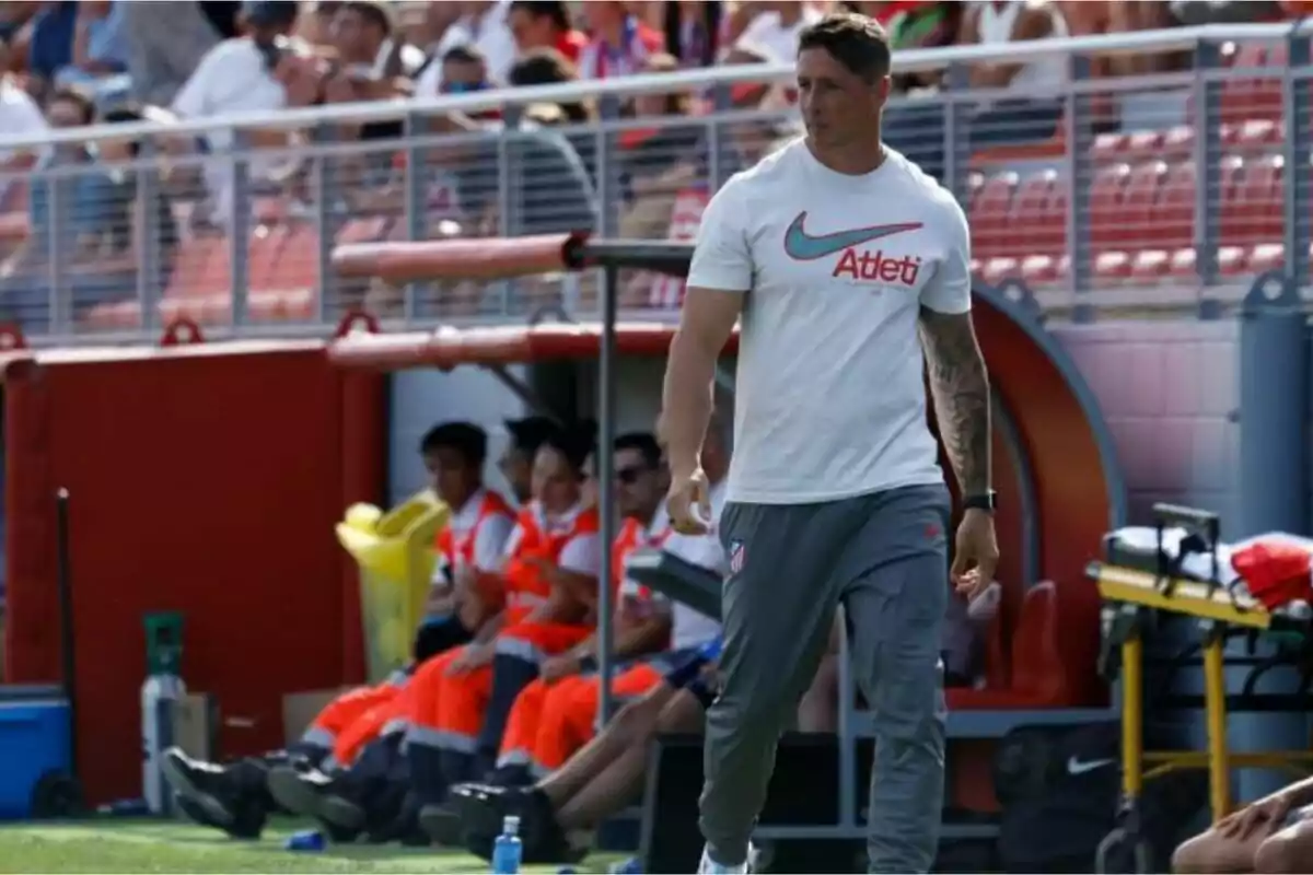 A man in a white T-shirt and grey sweatpants walks through the technical area of a football stadium as the public watches from the stands.