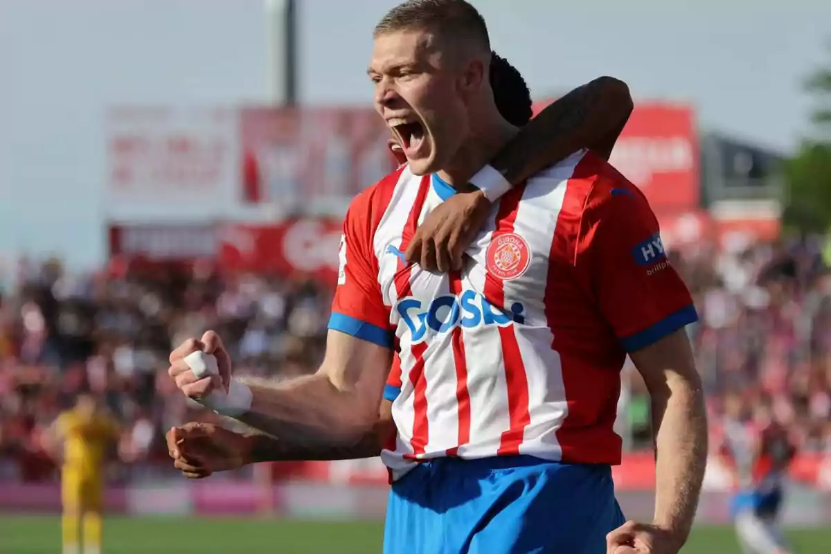 Un jugador de fútbol celebrando un gol con la camiseta del Girona FC mientras un compañero lo abraza.