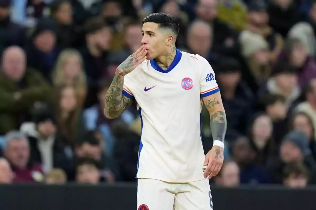 Un jugador de fútbol con uniforme blanco y detalles azules lleva su mano a la boca en un gesto durante un partido con espectadores desenfocados al fondo.