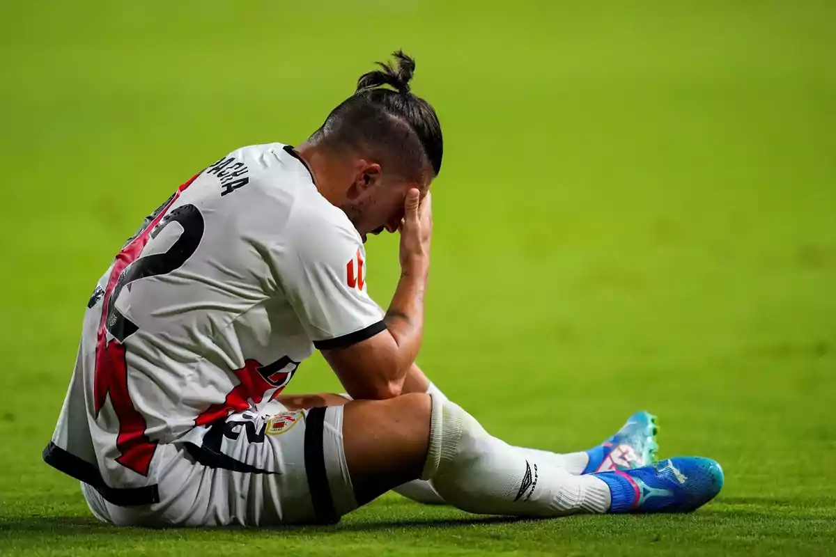 Jugador de fútbol sentado en el campo con la mano en la cara mostrando frustración.