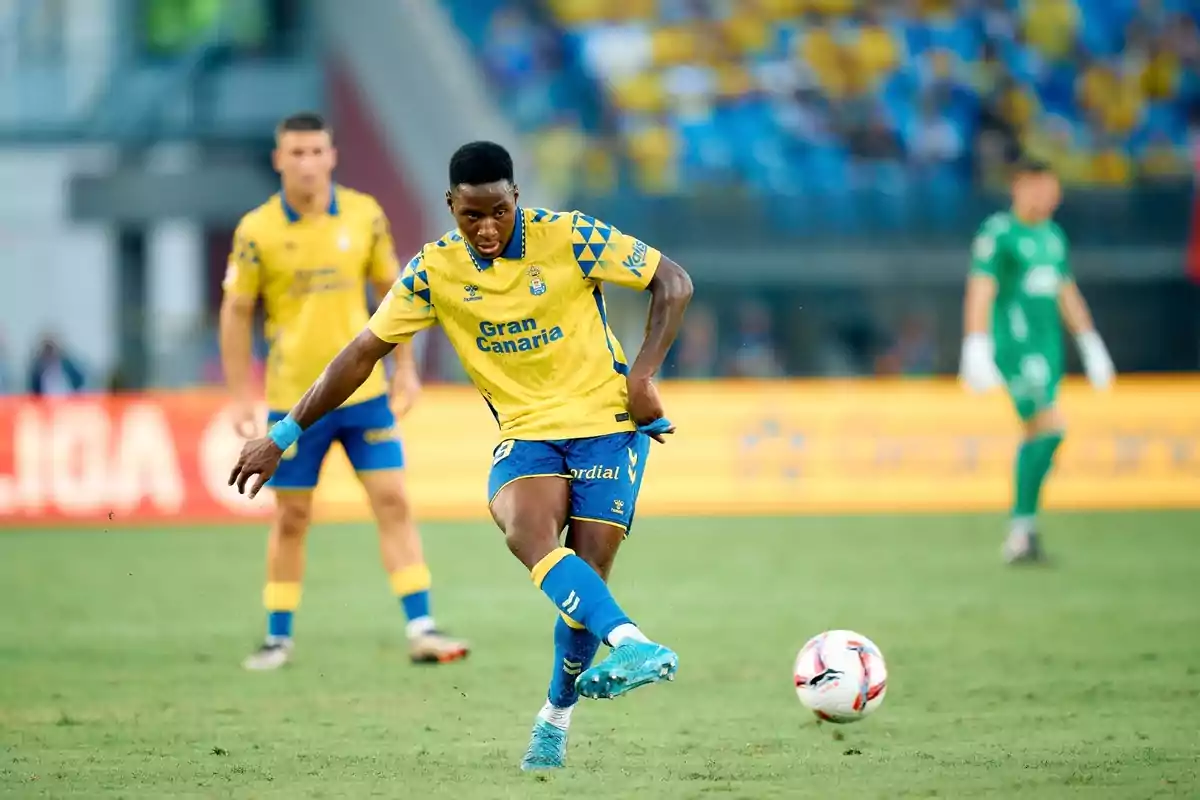 Jugador de fútbol con uniforme amarillo y azul pateando un balón en un estadio durante un partido.