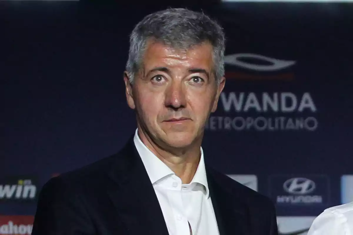 Un hombre de cabello canoso con traje oscuro y camisa blanca frente a un fondo con logotipos.