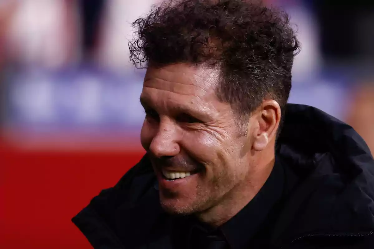 Hombre sonriendo con cabello rizado y barba ligera, vestido con chaqueta negra, en un entorno desenfocado.