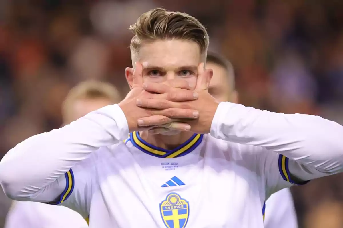 Un jugador de fútbol con uniforme blanco y detalles azules se cubre la boca con ambas manos.