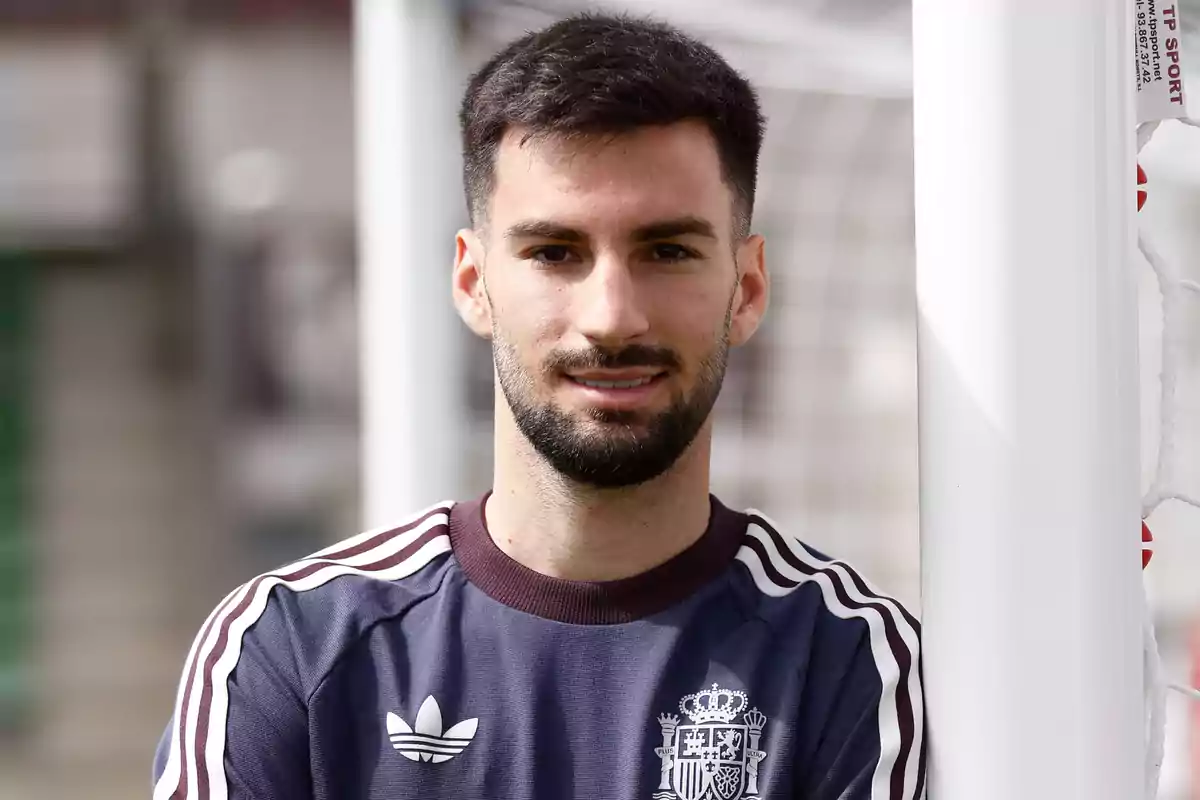 A man with a beard and short hair, wearing a sports sweatjersey with the Spanish coat of arms, is standing next to a soccer goal post.