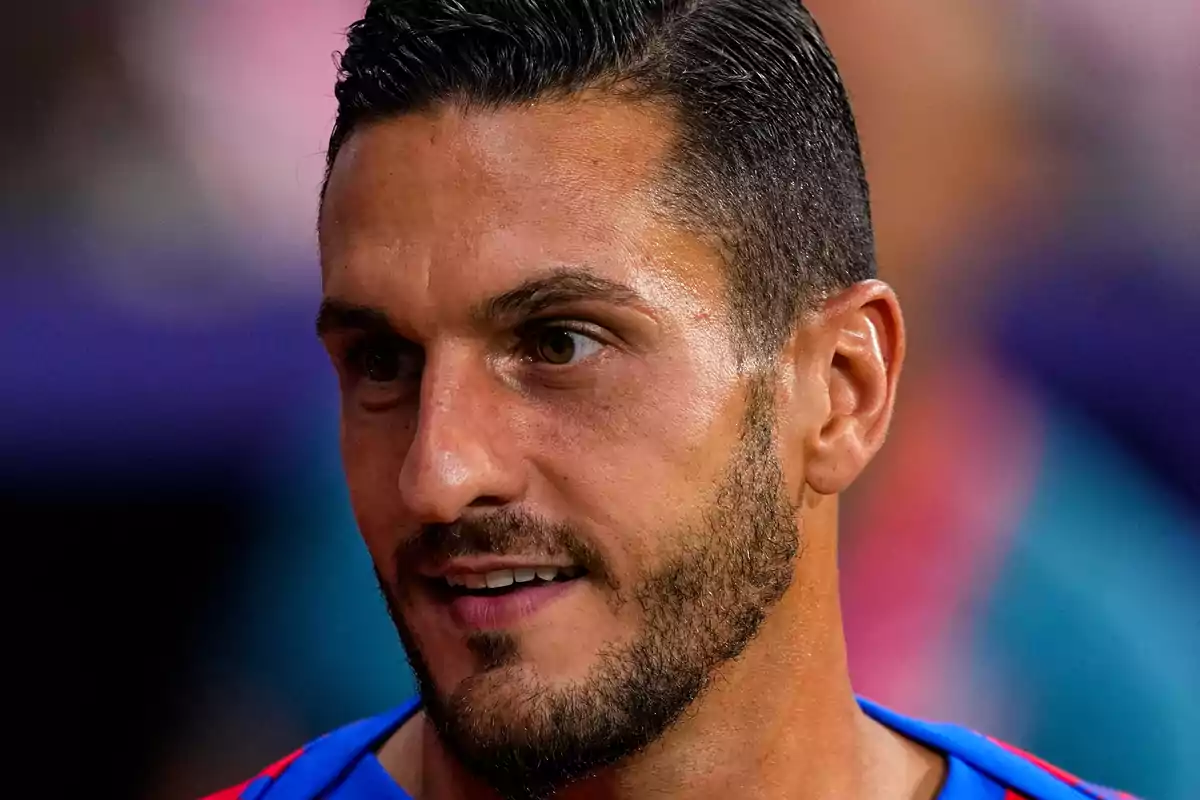 Un hombre con barba y cabello oscuro mira hacia un lado con una expresión neutral.