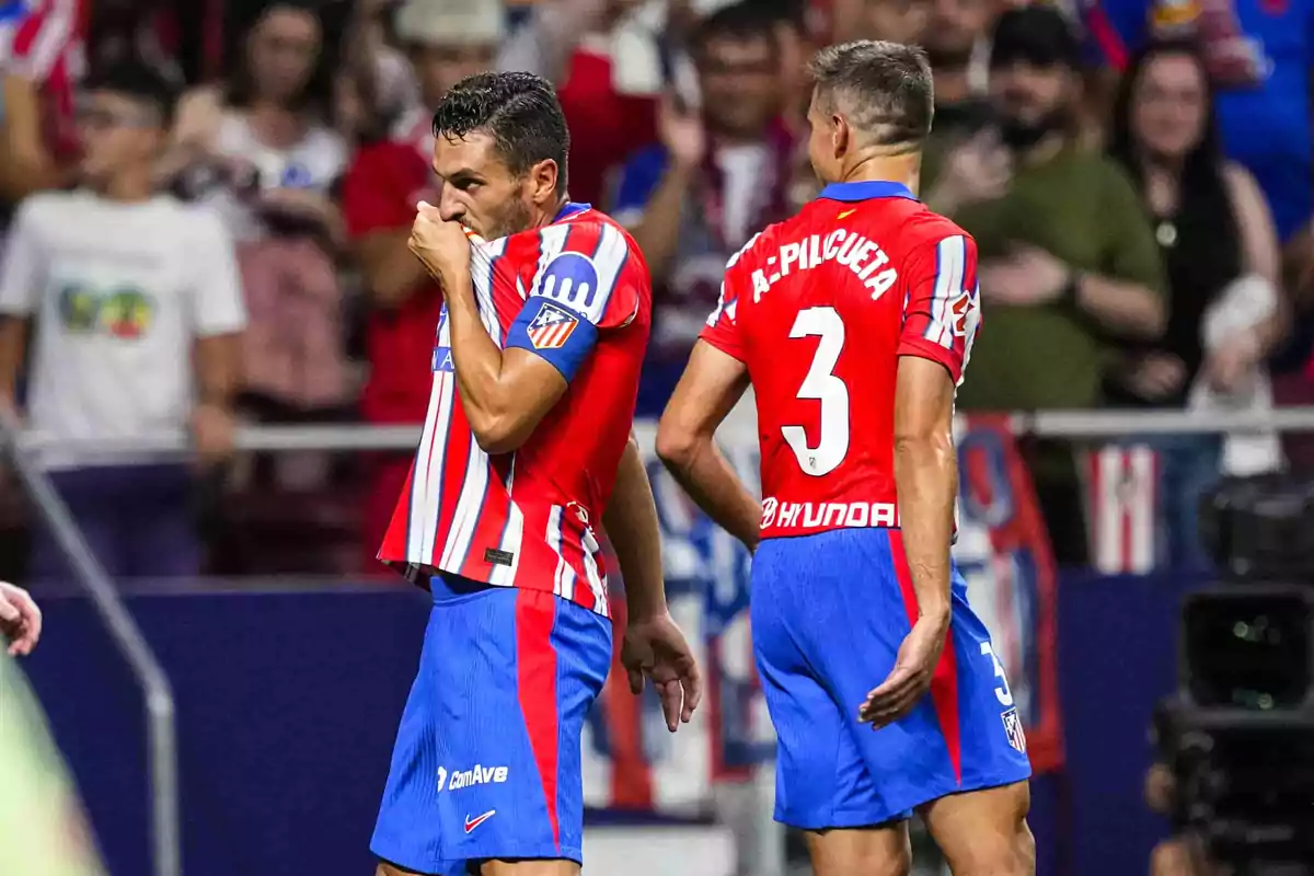 Dos jugadores de fútbol con uniforme rojo y azul en el campo, uno de ellos besa el escudo de su camiseta mientras el otro está de espaldas.