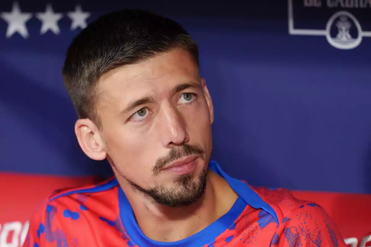 Un hombre con barba y bigote, vistiendo una camiseta roja con detalles azules, mira hacia un lado con una expresión seria.