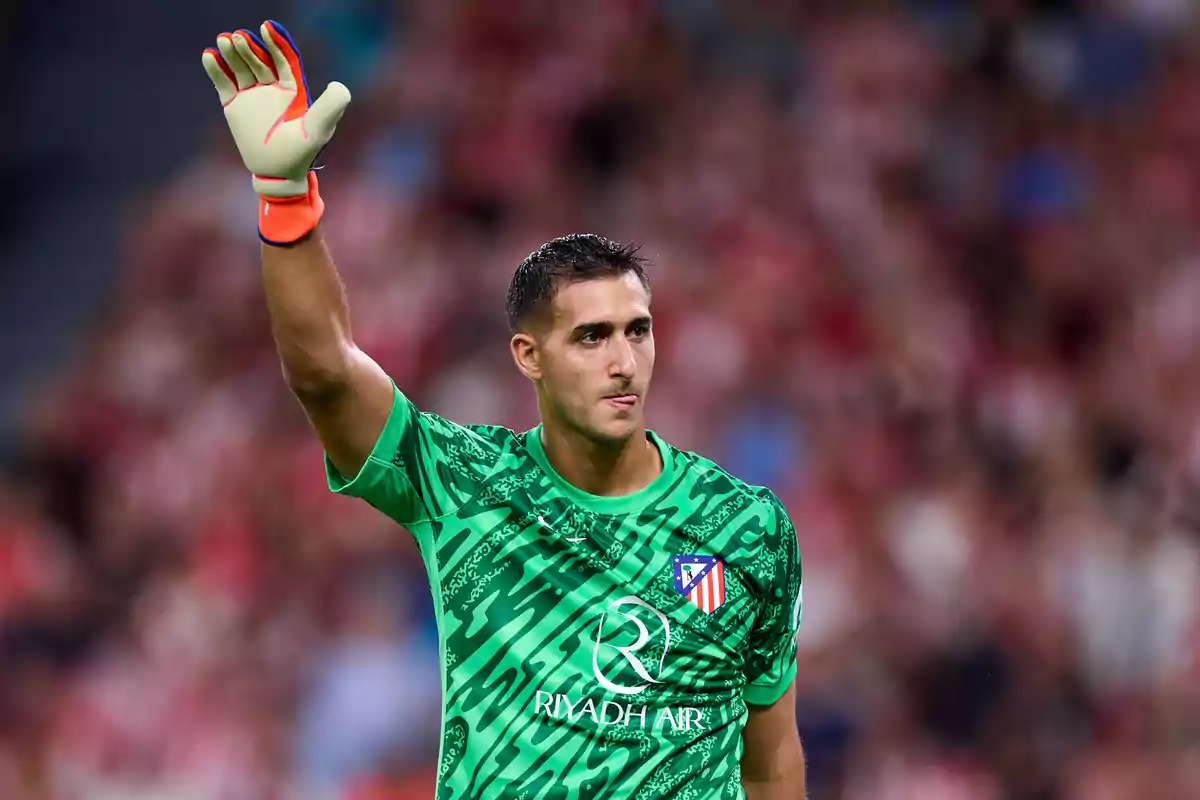 Portero de fútbol con uniforme verde levantando la mano en un estadio.