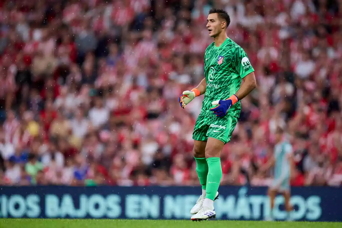 A football goalkeeper in a green uniform and colorful gloves stands in the background during a game with a lot of spectators.