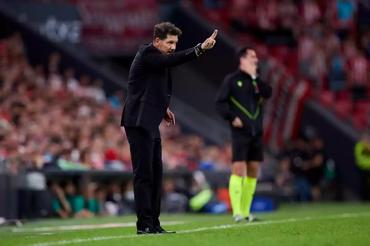 Un entrenador de fútbol vestido de traje negro da instrucciones desde la línea de banda durante un partido, con un árbitro asistente en el fondo.