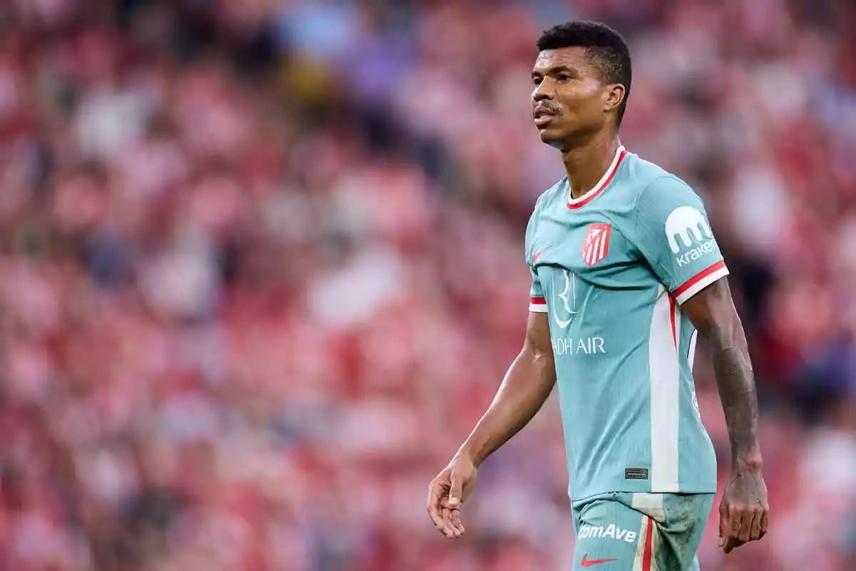 A soccer player in a light green uniform with red details, with a team logo on his chest, stands on the field with a blurred background of spectators.