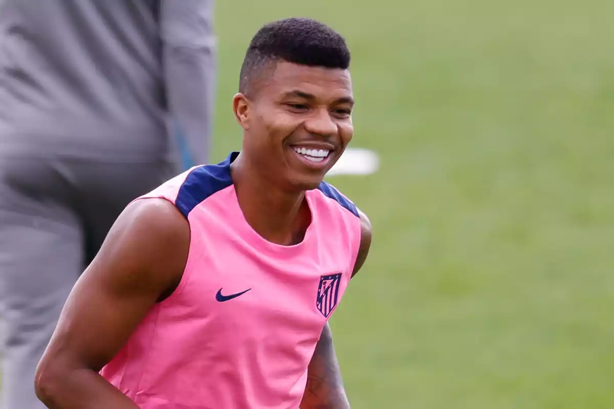 Un jugador de fútbol sonríe mientras entrena en el campo con una camiseta rosa.