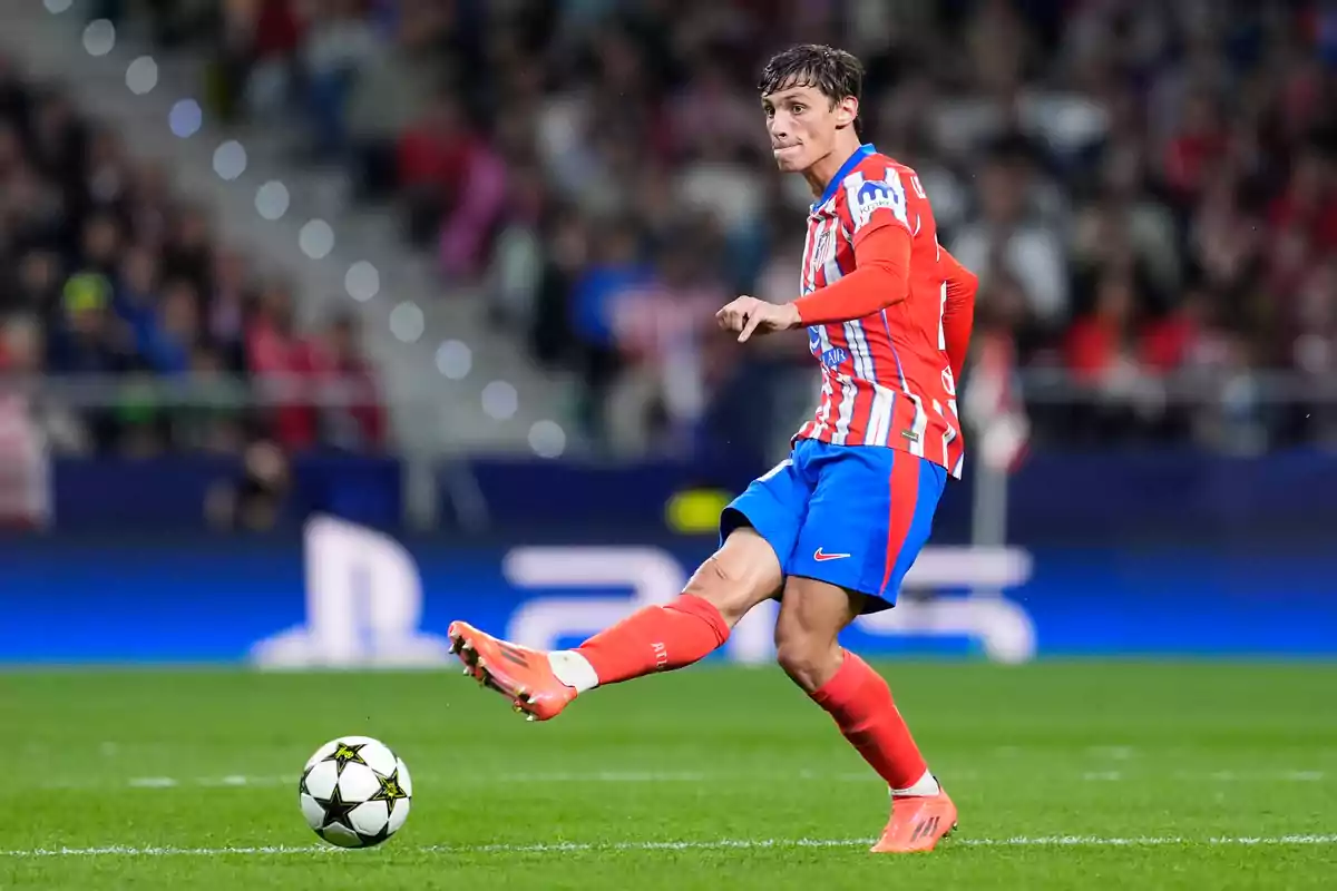 Jugador de fútbol con uniforme rojo y azul pateando un balón en un estadio durante un partido.