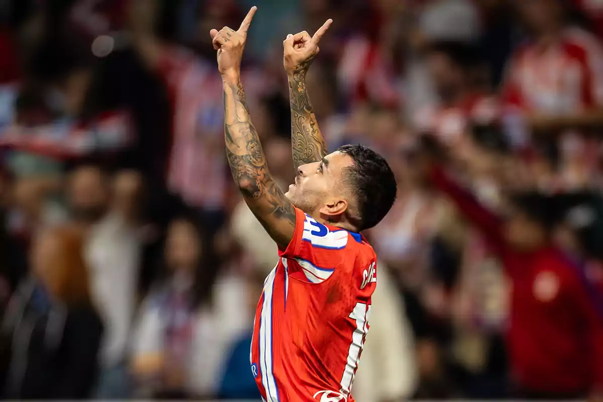 Jugador de fútbol celebrando con los brazos levantados y señalando hacia arriba, vistiendo un uniforme rojo con blanco y azul, con tatuajes visibles en ambos brazos y un estadio lleno de aficionados al fondo.