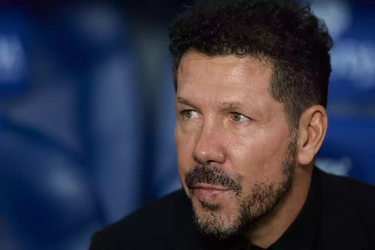 A man with a beard and curly hair looks to the side with a serious expression, with an out-of-focus background.