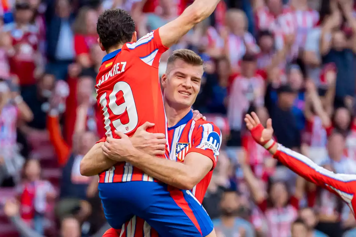 Dos jugadores de fútbol del Atlético de Madrid celebran un gol abrazándose en el campo, rodeados de aficionados.