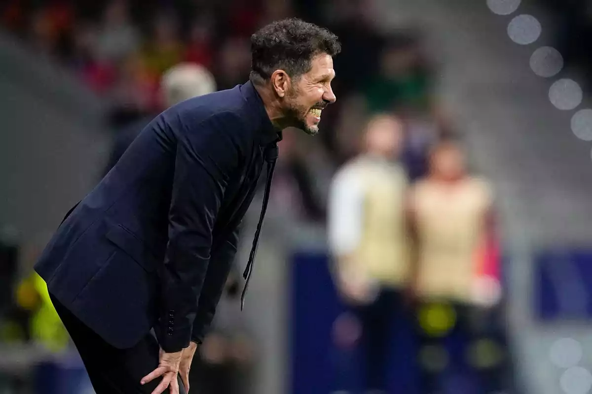 Un hombre con traje oscuro inclinado hacia adelante en un estadio de fútbol.