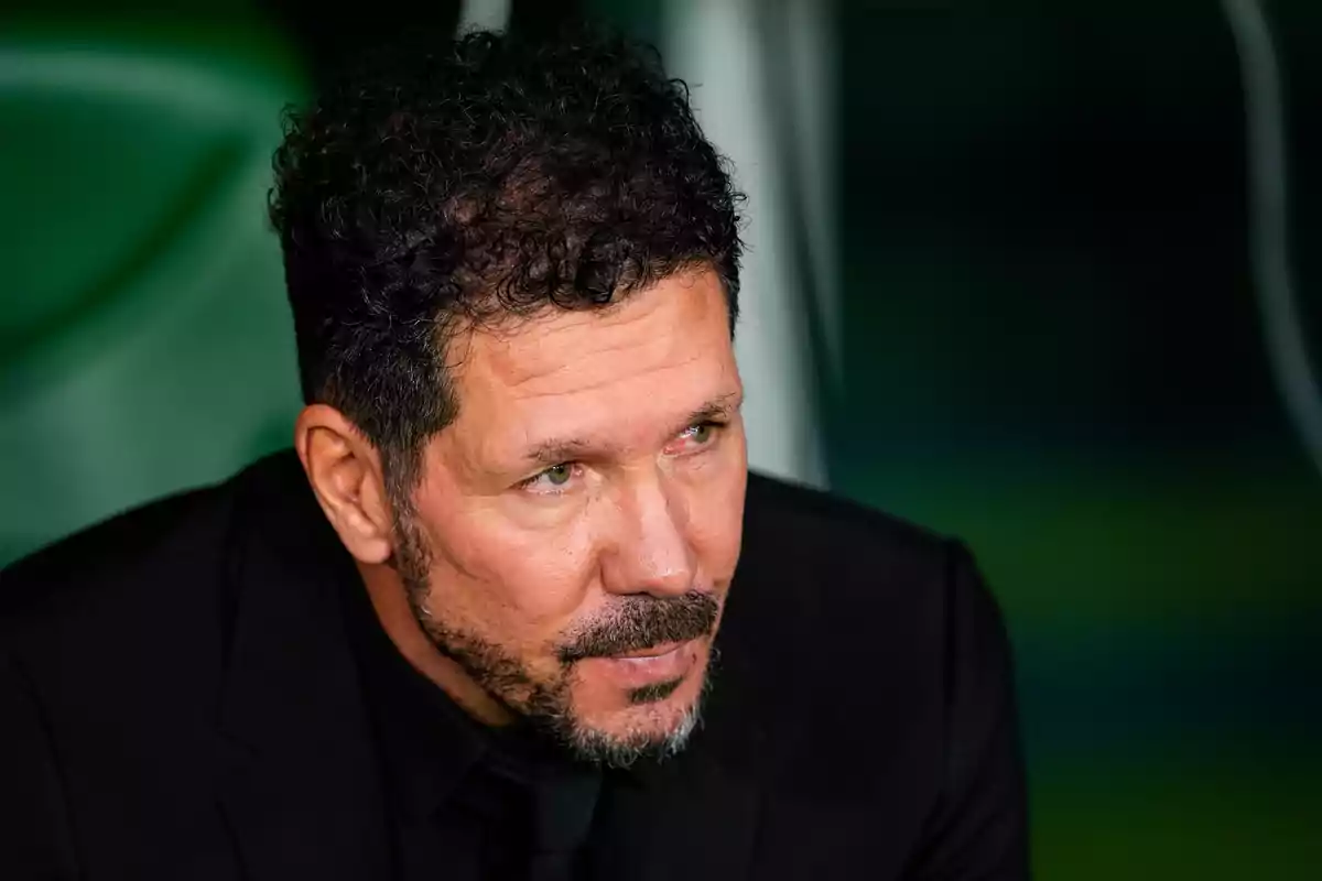 Hombre con cabello rizado y barba corta, vestido con traje negro, mirando hacia un lado con expresión seria.